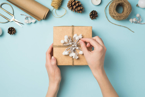 Première personne vue du dessus photo des mains de la femme décorant boîte cadeau avec brindille de neige et ficelle sur des boules d'arbre de Noël blanc et des outils d'artisanat sur fond bleu pastel isolé - Photo, image