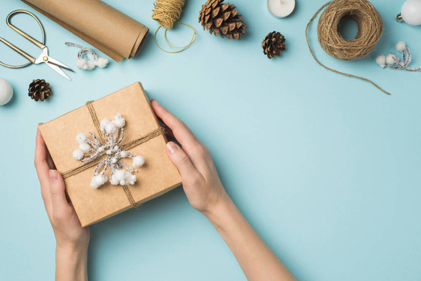 First person top view photo of hands holding kraft paper giftbox decorated with snow twig and twine christmas tree balls handicraft tools on isolated pastel blue background with blank space - Fotoğraf, Görsel