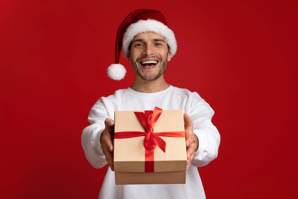 Geschenk für Sie. Aufgeregter junger Mann mit Nikolausmütze überreicht Weihnachtsgeschenk vor laufender Kamera - Foto, Bild