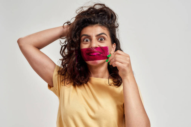 Amazed woman with curly hair holding purple film in front of her mouth, looking with eyes wide open at camera, adjusting hair posing isolated over light background - Foto, Imagem