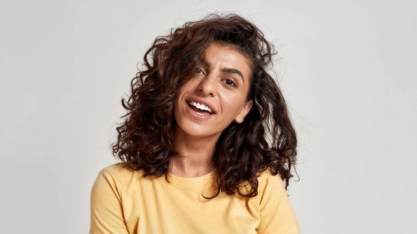 Closeup of pretty young woman with dark curly hair in casual wear looking cheerful at camera isolated over gray background - Foto, afbeelding