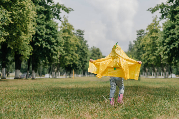 Happy little girl in yellow raincoat and muddy rubber boots running on dirt road through green grass near blooming rape seed field holding rhino in hand. Carefree childhood. - Foto, Bild