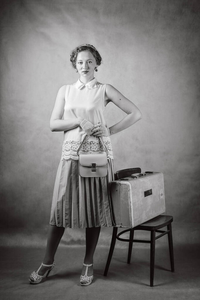 Young woman, dressed in retro style, with a handbag in her hands and a suitcase on a chair. Studio black and white portrait - Photo, Image