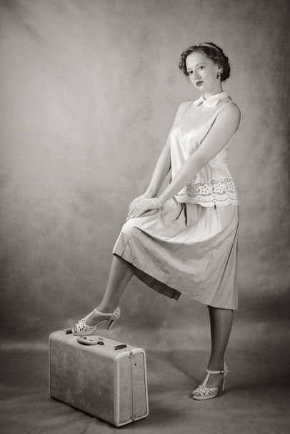 A young woman, dressed in retro style, stands with her foot on a suitcase. Studio black and white shot - Zdjęcie, obraz