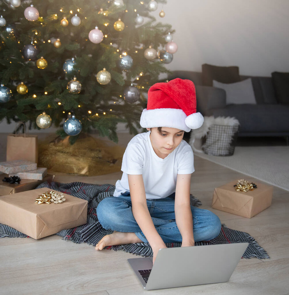 Chlapec sedí u vánočního stromku s laptopem. Vánoční stromek ve velkém obývacím pokoji. - Fotografie, Obrázek