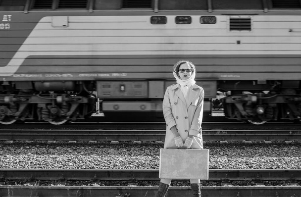 A young woman in a raincoat and dark glasses, with a suitcase in her hands, at the railway station. Retro style black and white outdoor shot - Zdjęcie, obraz