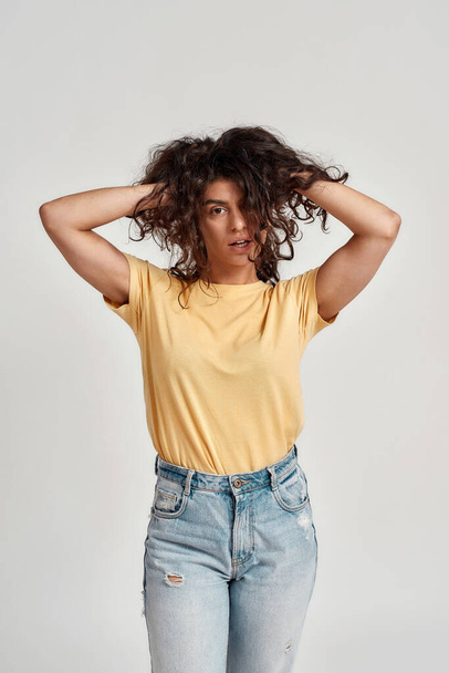 Playful young woman with dark curly hair in casual wear looking at camera, adjusting her hair while standing isolated over gray background - 写真・画像
