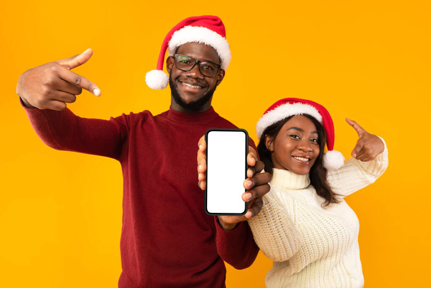 Африканська пара, яка демонструє смартфон на екрані, одягає Санта - Капелюхи, жовте тло - Фото, зображення