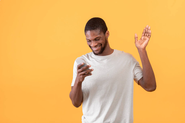 Happy african american man with smartphone making video call, waving hand at cellphone camera over yellow background - Photo, Image