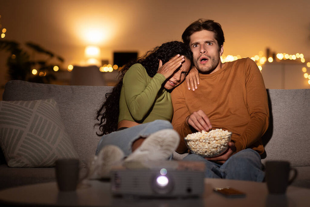 Σοκαρισμένο ζευγάρι βλέποντας ταινία τρόμου που καλύπτει τα μάτια χρησιμοποιώντας Projector Indoor - Φωτογραφία, εικόνα