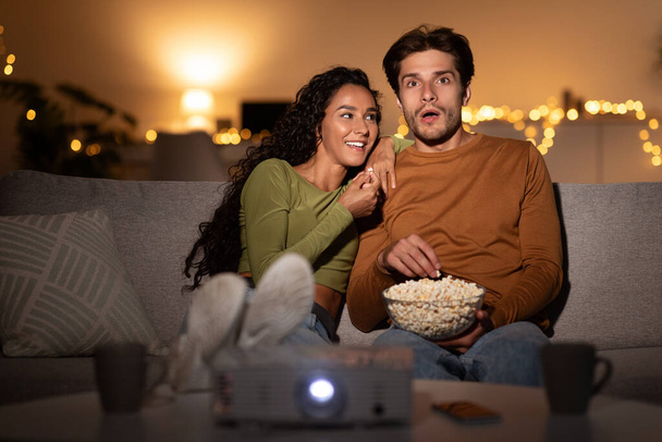 Emocionado pareja viendo película juntos usando proyector comiendo palomitas de maíz en interiores - Foto, Imagen