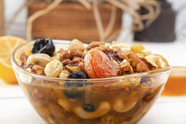 Суміш волоських горіхів, кешью, родзинок, сушених абрикосів і чорносливу з медом у склянці на білому дерев'яному столі. Крупним планом
 - Фото, зображення