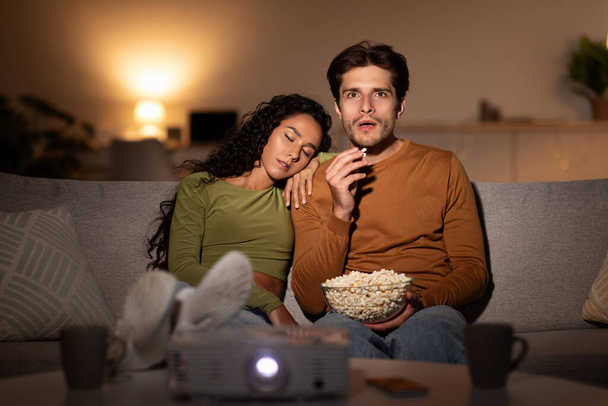 Femme qui dort pendant que son mari regarde un film en utilisant un projecteur à la maison - Photo, image