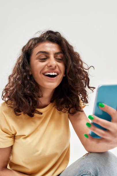Giovane donna gioiosa con i capelli ricci scuri sorridente mentre fa videochiamata utilizzando smartphone isolato su sfondo chiaro - Foto, immagini