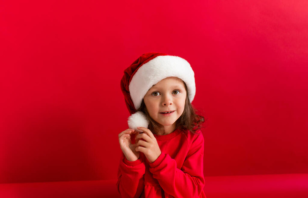Cute little girl in a red Christmas hat and red dress on a red background. Merry Christmas and Happy Holidays. - Foto, afbeelding
