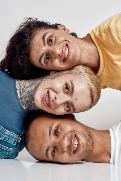 Detailní portrét tří veselých různorodých přátel usmívajících se na kameru, ležící hlavou na sobě, pózující izolované nad světlým pozadím - Fotografie, Obrázek