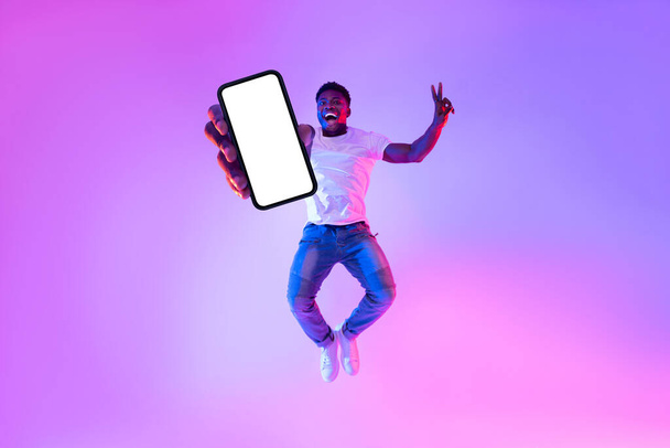 Täysi pituus innoissaan Afrikkalainen amerikkalainen kaveri hyppäämällä älypuhelimella, osoittaa tyhjä näyttö neon valossa, mockup - Valokuva, kuva
