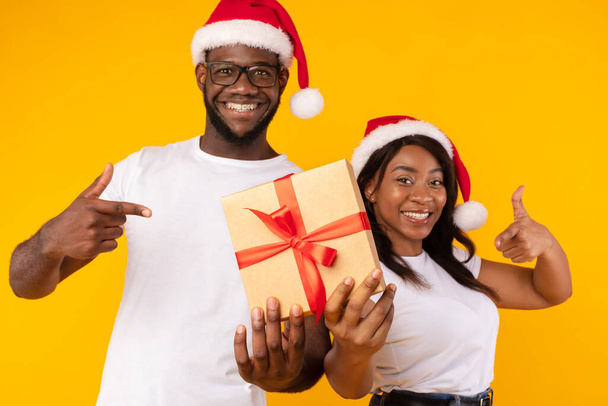 Afrikanisches Paar hält Weihnachtsgeschenk in der Hand und gestikuliert mit Daumen nach oben über gelbem Hintergrund - Foto, Bild
