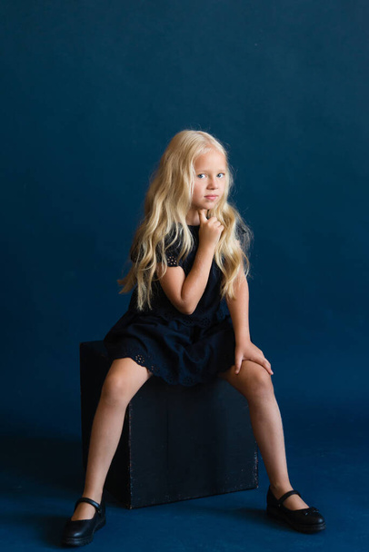 beautiful blonde girl in a dark blue dress on a solid blue background. The photo is great for demonstrating the character of a child - daydreaming, enthusiasm, vulnerability, touching, beauty, stubbornness. - Foto, Imagen