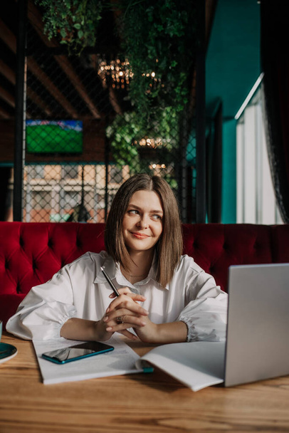 uma mulher de negócios morena sorridente em uma camisa branca está sentada a uma mesa em um café com um laptop - Foto, Imagem