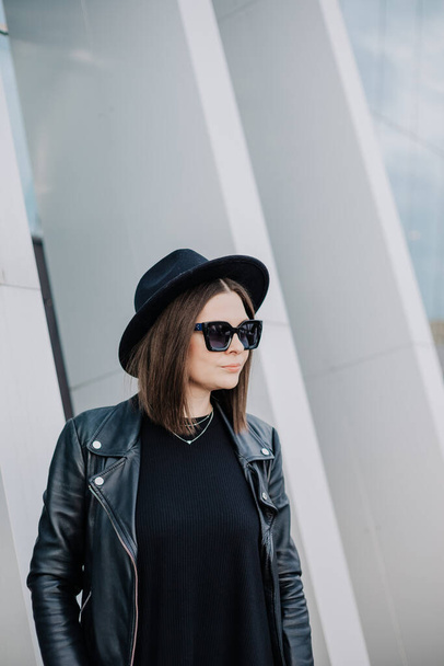 una mujer joven en un vestido negro y una chaqueta de cuero con gafas negras está de pie en la calle - Foto, Imagen