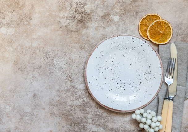 Winter holiday table setting. Craft ceramic plate, linen napkin, cutlery and dry oranges slices. Festive composition. Flat lay. - Fotografie, Obrázek