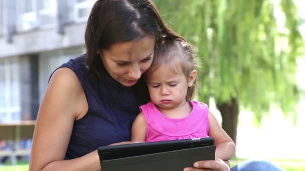 Madre enseña a una niña pequeña.Mamá enseña a un niño a jugar en la tableta.Madre joven con el bebé en el parque jugando en la computadora portátil
. - Metraje, vídeo