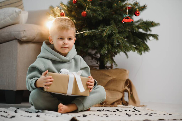 The boy sits near the Christmas tree with gifts. Children for the Christmas holidays. - Foto, Imagen