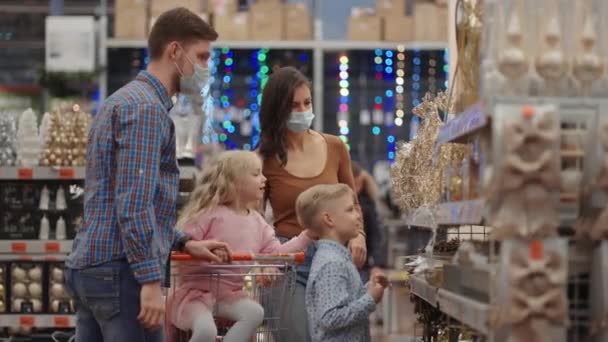 Vier mensen, papa en twee kinderen met een winkelwagentje. Een gelukkig gezin in medische maskers in de winkel koopt kerstversieringen en geschenken in slow motion - Video