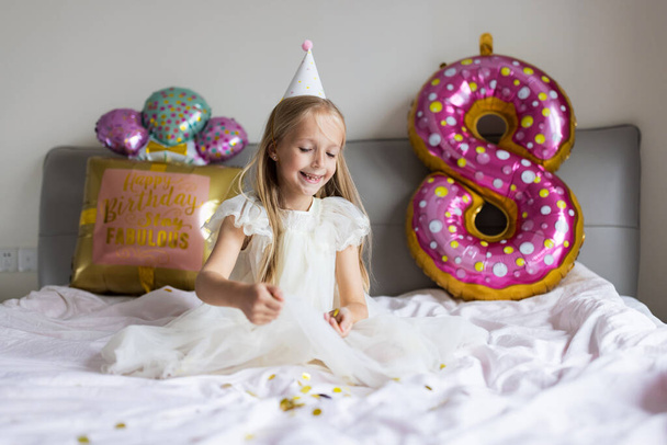 Indoor shot of pretty joyful cute adorable girl kid celebrating eight years old birthday with bright and colorful balloons with inscription words stay fabulous, wearing casual fashionable dress - Photo, image