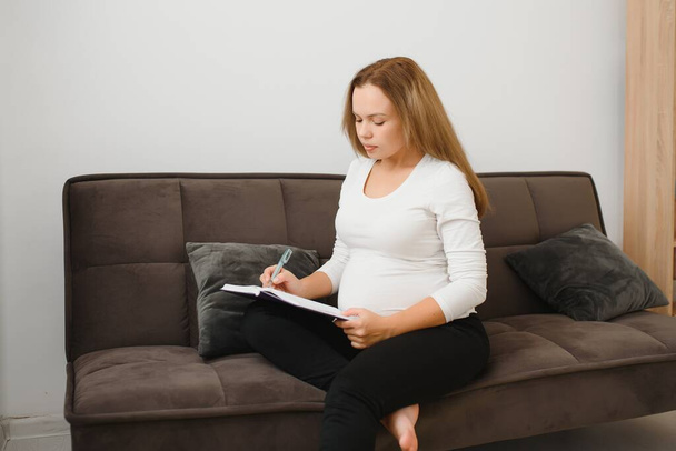 Pregnant woman lying on the sofa and making notes in her diary she resting at home - 写真・画像