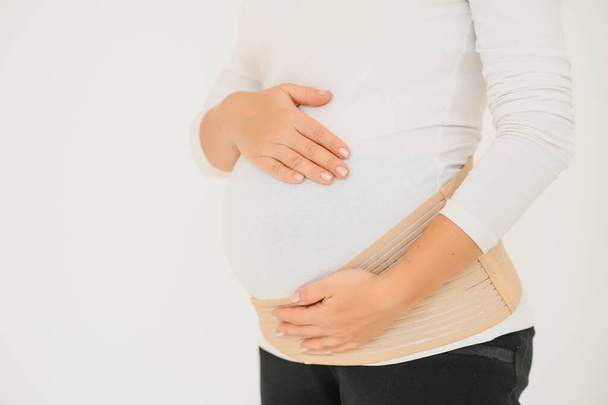 Close up of pregnant woman putting on a bandage at gray background with copy space. Orthopedic abdominal support belt concept. - Foto, imagen