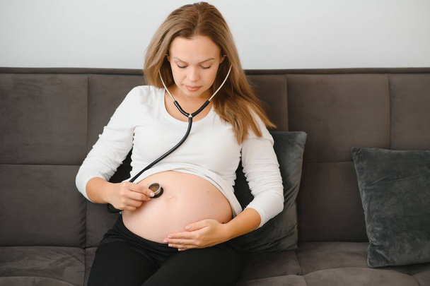 The mother is using stethoscope the doctor listens to the sound of the baby in the stomach. Asian pregnant woman using stethoscope listening her baby on sofa. - Фото, зображення