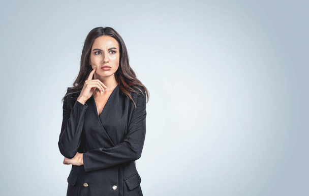 Thoughtful young attractive businesswoman wearing formal suit is standing in front of the wall touching her chin. Concept of imagination and inspiration for creative ideas - Foto, Bild