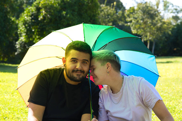 persona de género no binario y un hombre gay están sentados en el parque, cubriéndose del sol con un paraguas orgullo gay. están posando para la cámara y muy felices. Diversidad y orgullo gay. - Foto, imagen