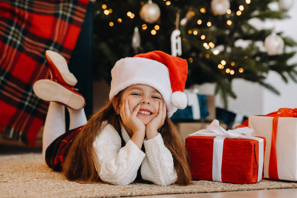 A happy little girl in a Santa Claus hat smiles with gifts in her hands. - Foto, afbeelding