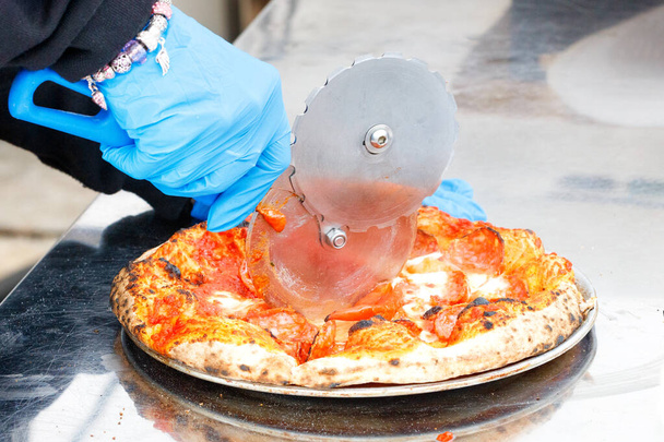 De handen van de ober in blauwe latex handschoenen snijden de gebakken pizza met een rond metalen mes. Kopieerruimte. - Foto, afbeelding