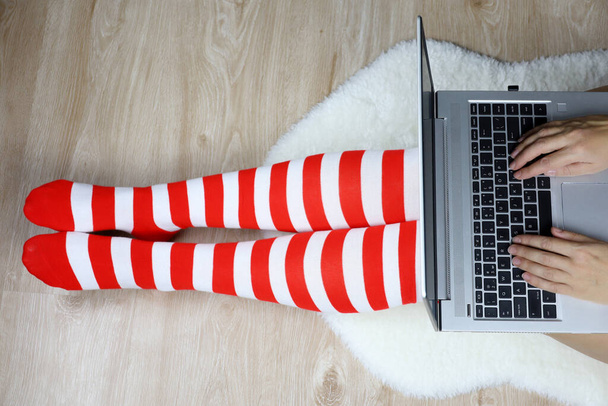 Woman in Christmas knee socks sits on a fur rug with laptop, top view. Businesswoman, work and leisure at home during New Year holidays - Foto, afbeelding