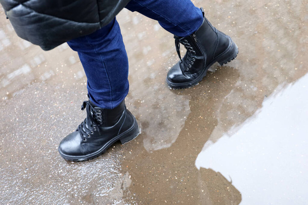 Rain in city, female legs in boots on a street with puddles. Residential buildings are reflected in water, woman walking in autumn or winter weather - Photo, Image