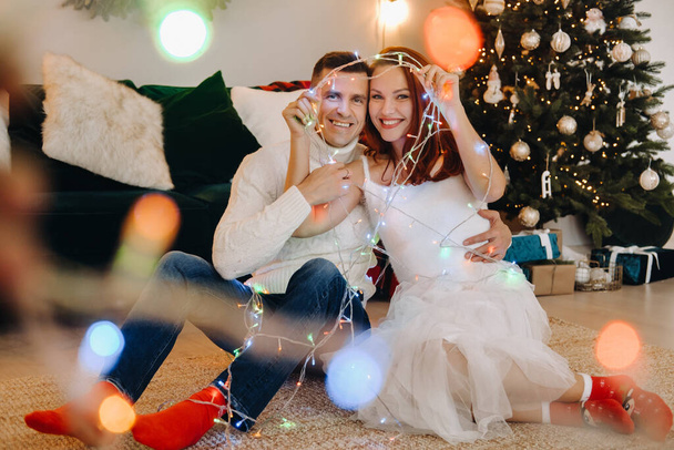 Ein glückliches Ehepaar sitzt zu Hause auf dem Fußboden neben dem Weihnachtsbaum. - Foto, Bild