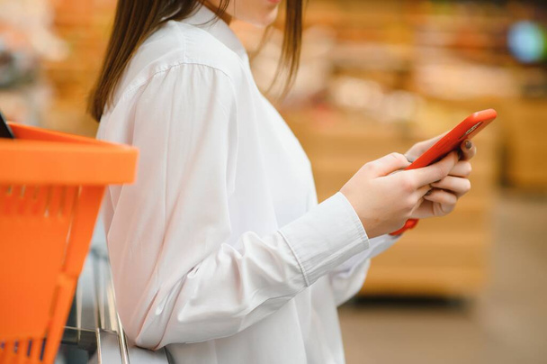 Woman doing grocery shopping at the supermarket, she is leaning on the shopping cart and connecting with her phone, apps and retail concept - 写真・画像