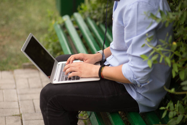 Image fermée d'un étudiant utilisant un ordinateur portable et écoutant une musique dans un casque, assis sur un banc dans un parc urbain. Concept d'éducation et de loisirs. - Photo, image