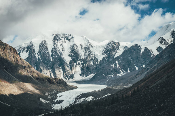 Beautiful mountain landscape with glacier tongue and great snowy mountains in sunlight in cloudy sky. Scenic alpine landscape with glacial tongue and high mountains with glaciers in low clouds. - Photo, Image