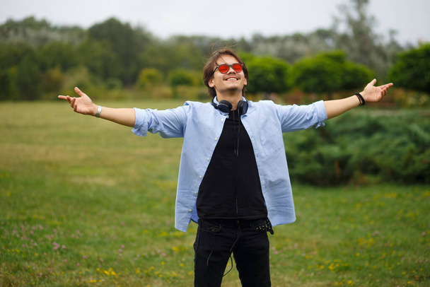Happy youth boy with raised hands and red sunglasses sitting in park, feelings happiness. Freedom, emotional concept. - Photo, Image