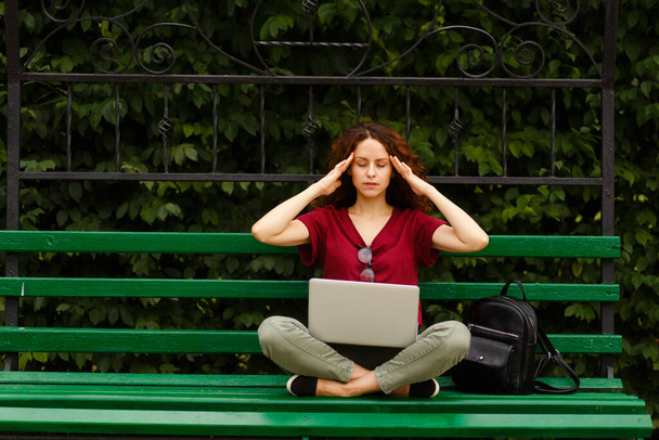 Кучерява молода жінка з закритими очима, працює на ноутбуці, сидить на зеленій лавці в парку гастролює її храмами
. - Фото, зображення