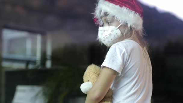 Menina de seis anos de idade no chapéu de Papai Noel senta-se na mesa sozinho com ursinho de pelúcia. - Filmagem, Vídeo