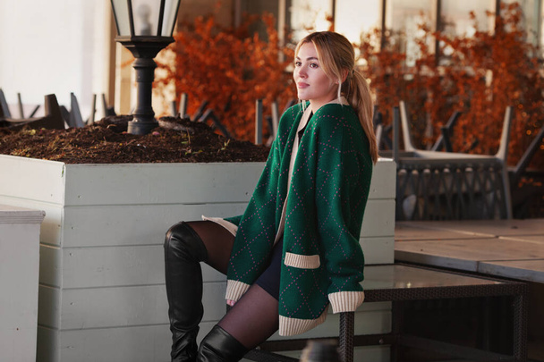 Mujer hermosa pensando o esperando. Elegante modelo de moda con suéter verde de gran tamaño, pantalones cortos, mirando hacia otro lado. Fondo hojas de otoño y campo de profundidad - Foto, imagen