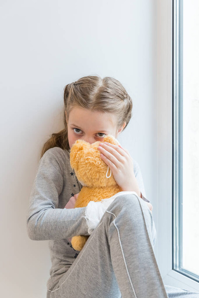 портрет грустного ребенка, сидящего на подоконнике с мягкой игрушкой. Больной ребенок с жаром и болезнями - Фото, изображение