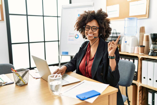 African american woman with afro hair working at the office wearing operator headset with a big smile on face, pointing with hand finger to the side looking at the camera.  - Fotó, kép