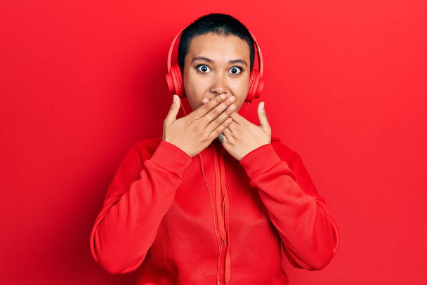 Beautiful hispanic woman with short hair listening to music using headphones shocked covering mouth with hands for mistake. secret concept.  - Photo, Image